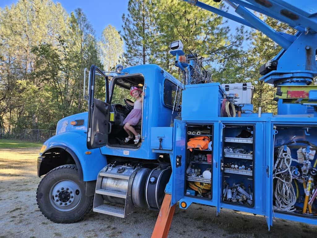 Blue fire truck with a young girl in a pink dress and helmet sitting in the driver's seat on a school campus.