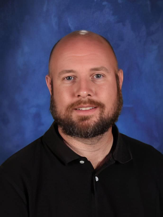 Bald man with a beard wearing a black polo shirt against a blue background at El Dorado Adventist School.